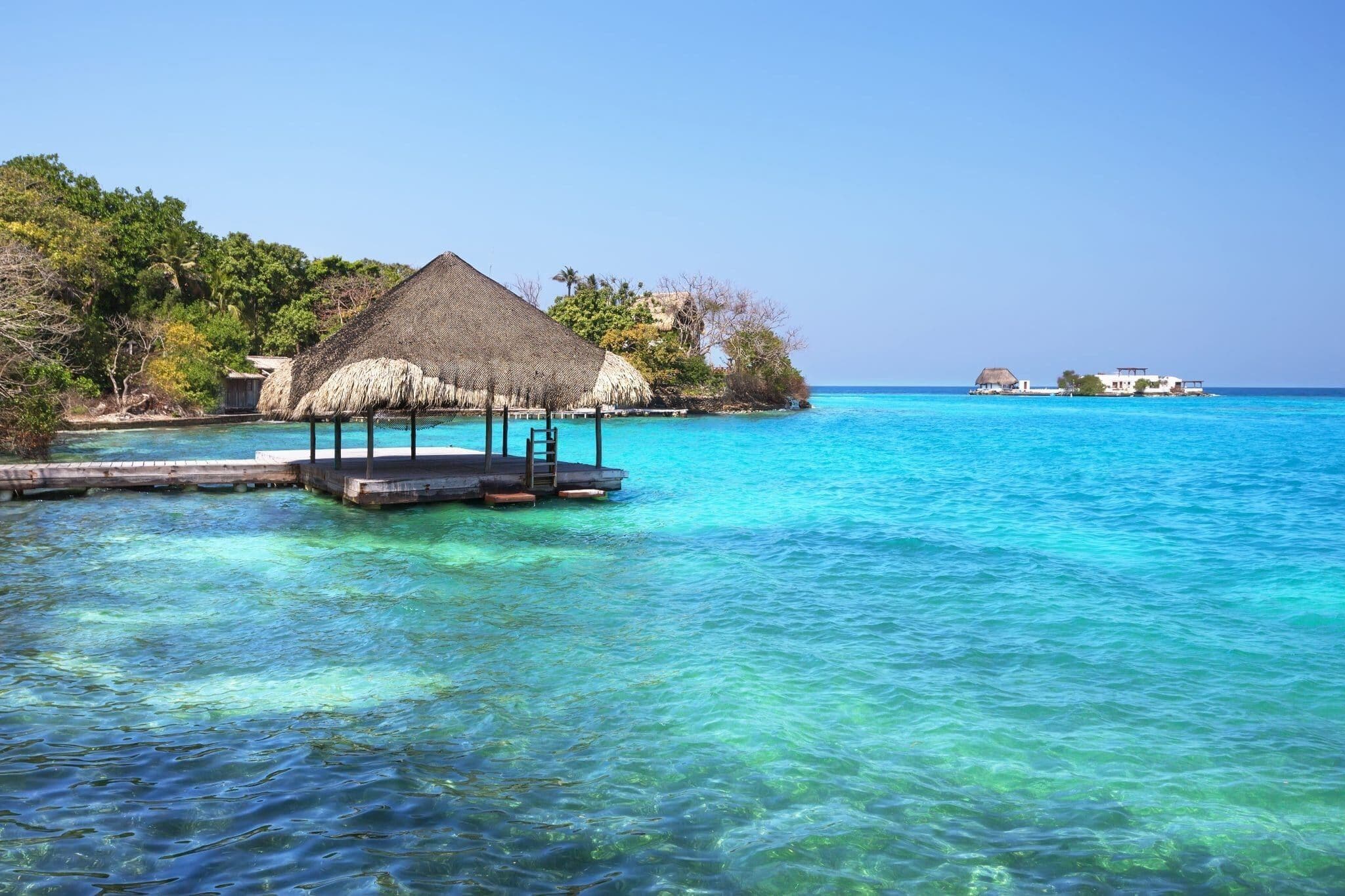 A clear turquoise sea with a wooden dock leading to a thatched-roof structure.