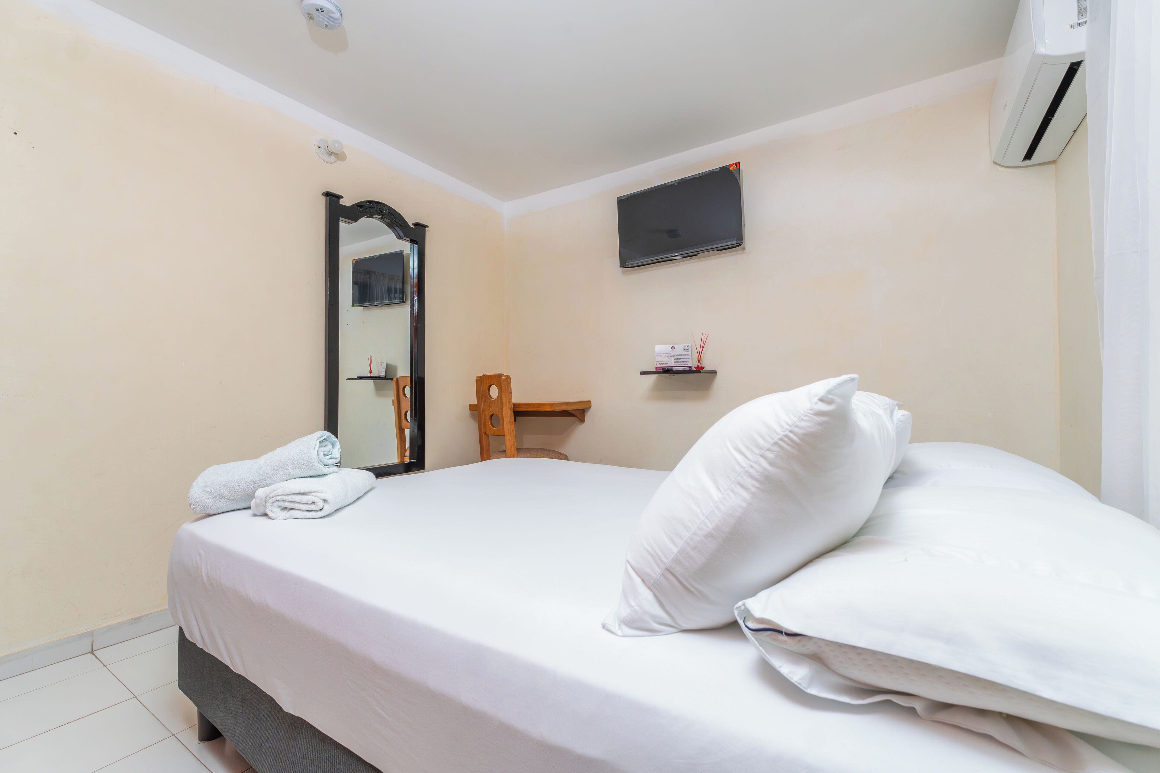 A neatly made bed with white linens and pillows, a flat-screen TV mounted on the wall, and a small shelf with toiletries.