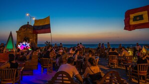 Un espacio al aire libre con sillas y mesas de madera, un escenario con un letrero de neón y banderas.