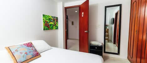 Una habitación de hotel con cama, mesita de noche, espejo, armario y puerta que da a otra estancia.