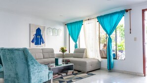 A living room with a sofa, a teal armchair, a coffee table with books, and a window with curtains.