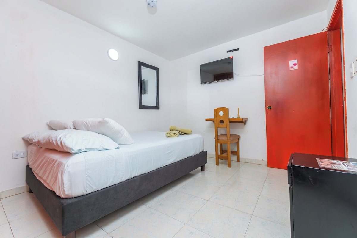 A bedroom with a bed, a desk, a chair, a red door, and a TV on the wall.