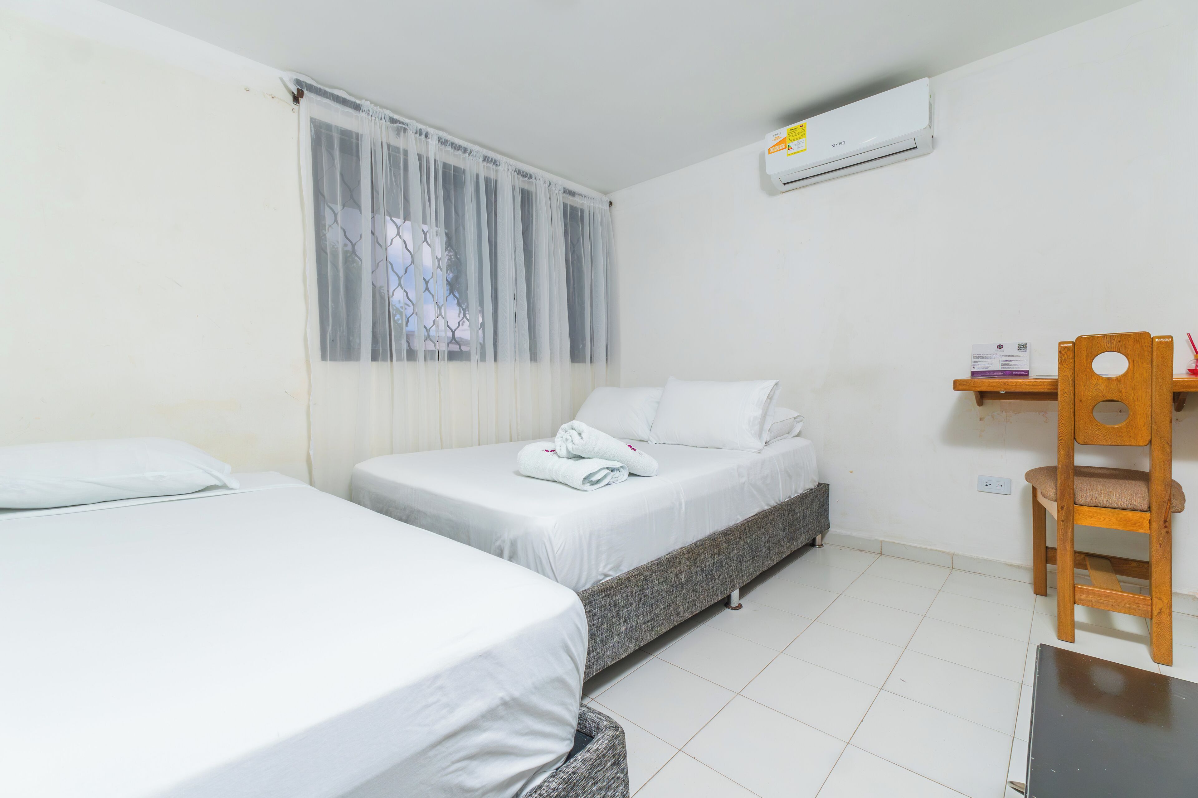 Une chambre d’hôtel avec deux lits simples, une chaise en bois, un petit bureau et un appareil de climatisation fixé au mur.