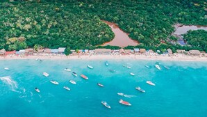 Vista aérea de una playa con numerosos barcos en el agua y una densa zona boscosa al fondo.
