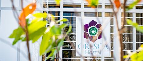 Een bord met de tekst 'CRESCA Hotel' en een logo, deels verborgen achter een metalen hek en begroeiing.