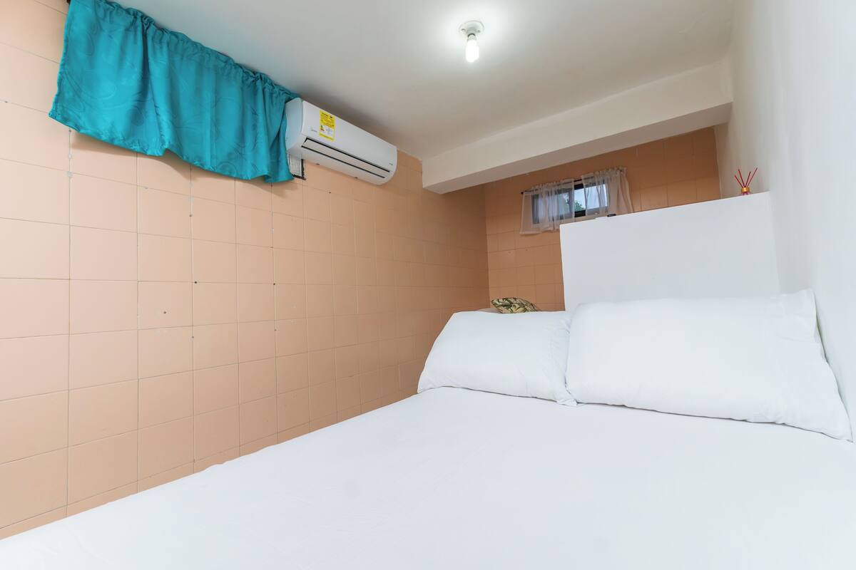 Une chambre à coucher avec un lit, des oreillers blancs, un rideau turquoise, un appareil de climatisation et une fenêtre avec un rideau.