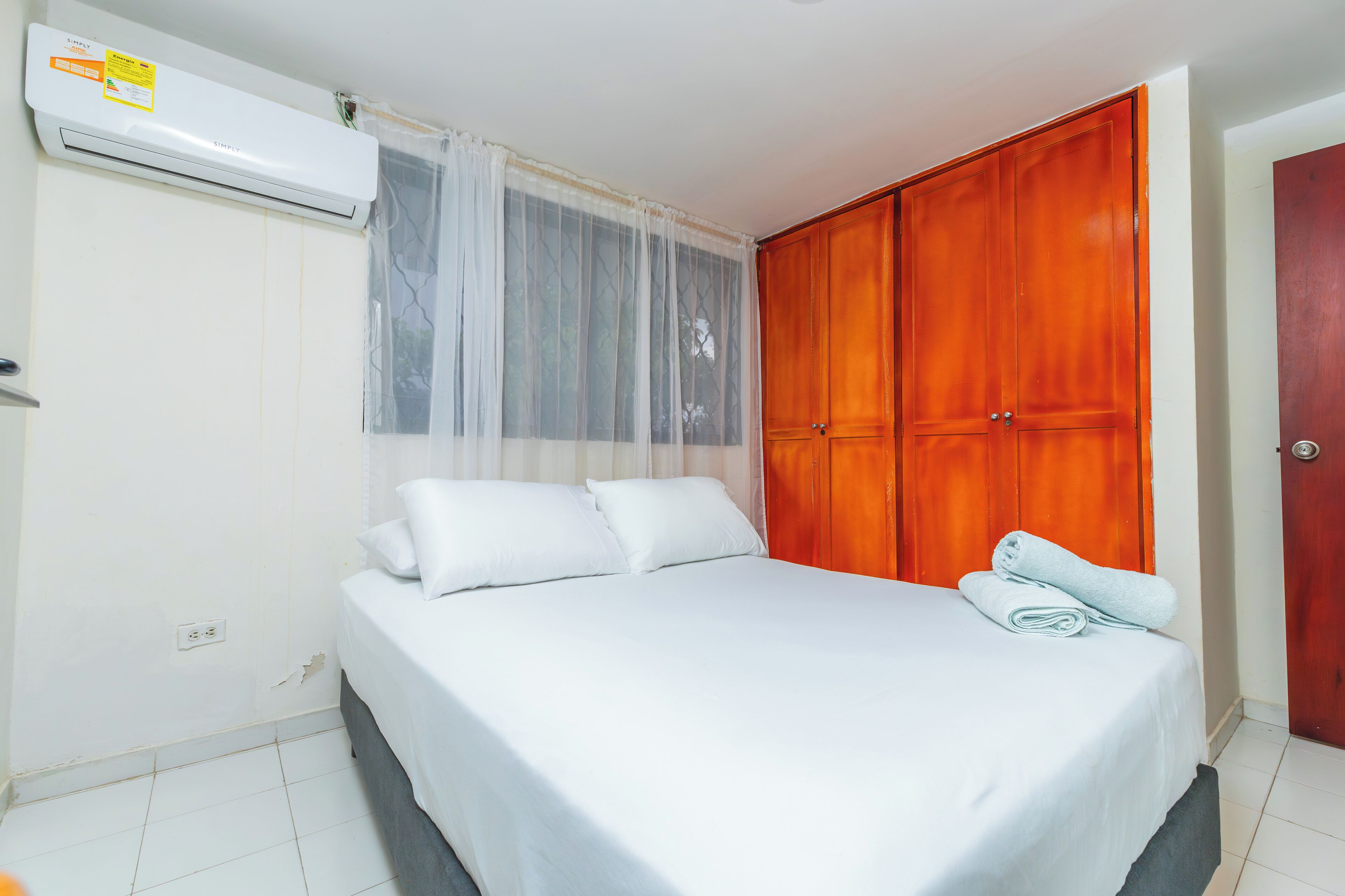 A bedroom with a bed, white linens, a wooden wardrobe, and an air conditioning unit.