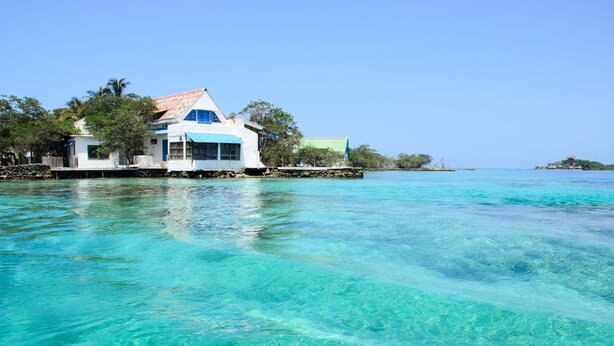Una propiedad frente al mar con una casa sobre pilotes, elevada sobre aguas turquesas cristalinas, rodeada de árboles y un cielo despejado.