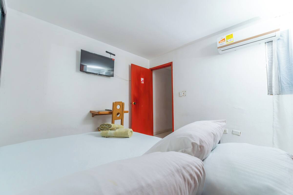 Une chambre d’hôtel avec un lit, une porte rouge, un téléviseur fixé au mur et un appareil de climatisation.