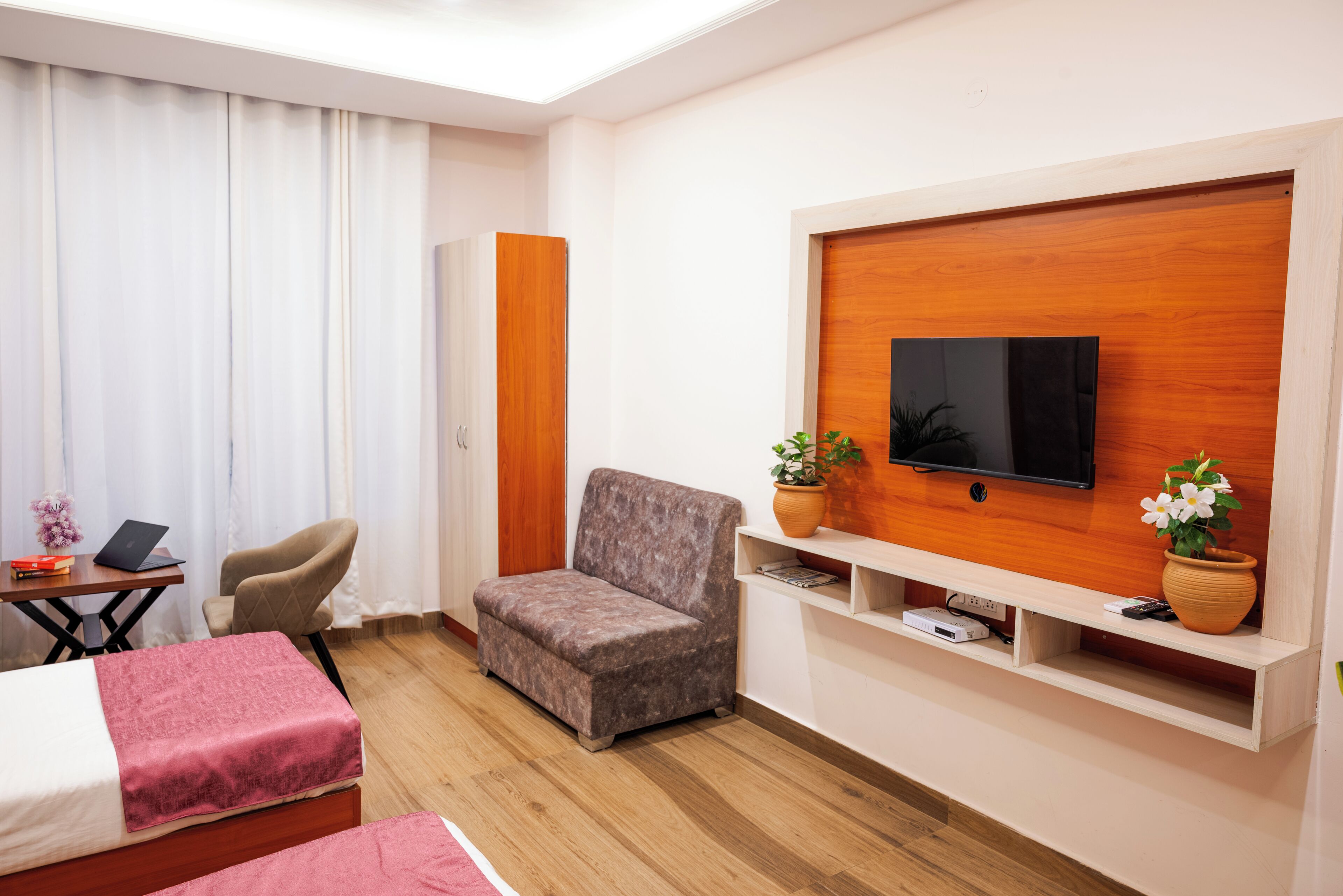 Une chambre d’hôtel avec un lit, un bureau avec un ordinateur portable, une chaise, un téléviseur fixé à un mur en bois et deux plantes en pot.