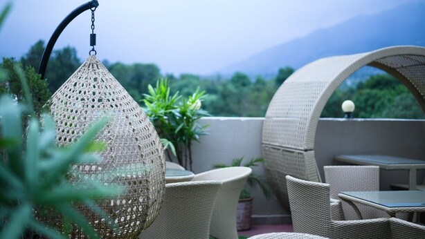 A wicker outdoor seating area with tables and chairs, a hanging light fixture, and a view of greenery and mountains in the background. - Hotel Sagar Residency- Best Mountain View Hotel In Palampur (Palampur)