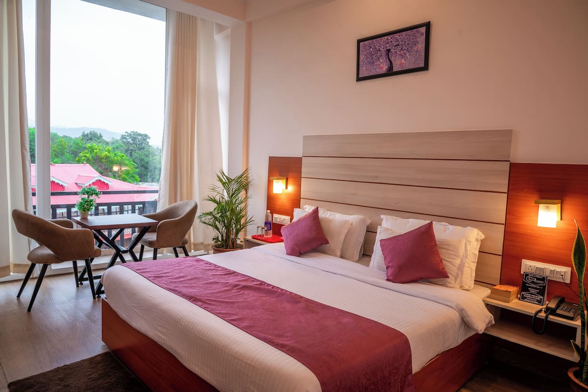 A hotel room with a large bed, two bedside tables, a desk with chairs, a potted plant, and a view of a red-roofed building.