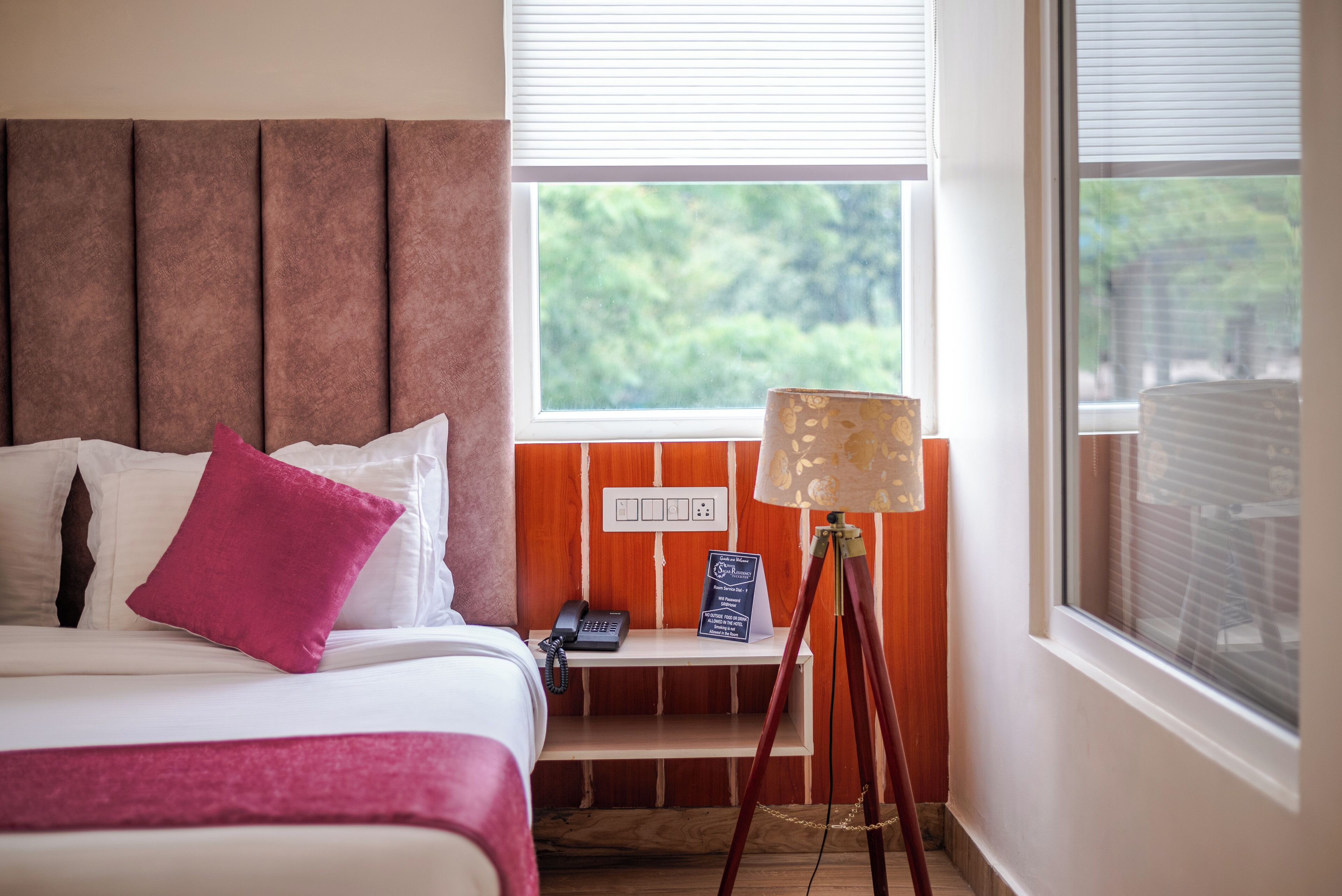 A hotel room with a bed, a bedside table with a lamp, a phone, and a window with a view of greenery.