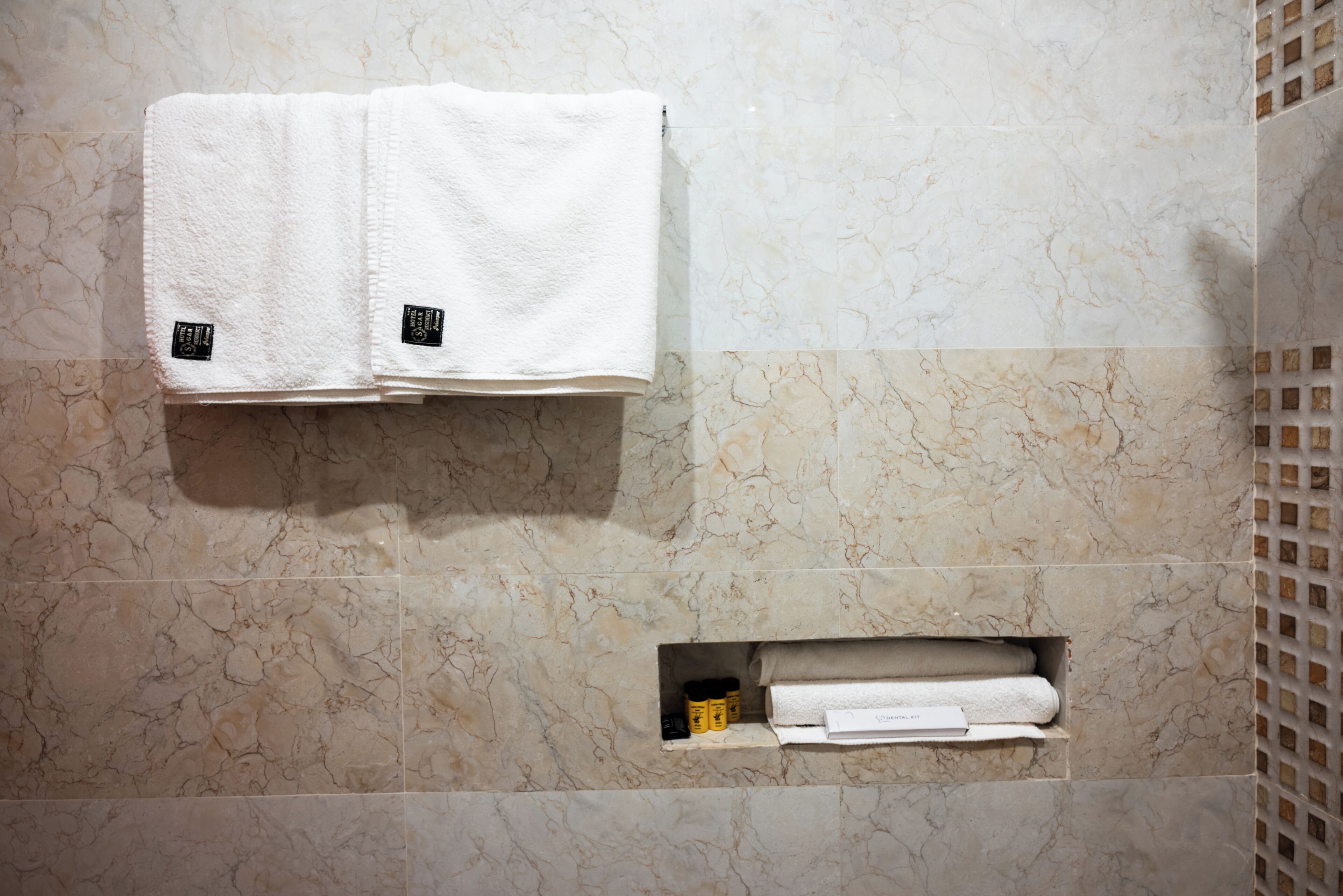 Un baño con baldosas de mármol, dos toallas colgadas en un perchero, y un estante con toallas y artículos de aseo adicionales.