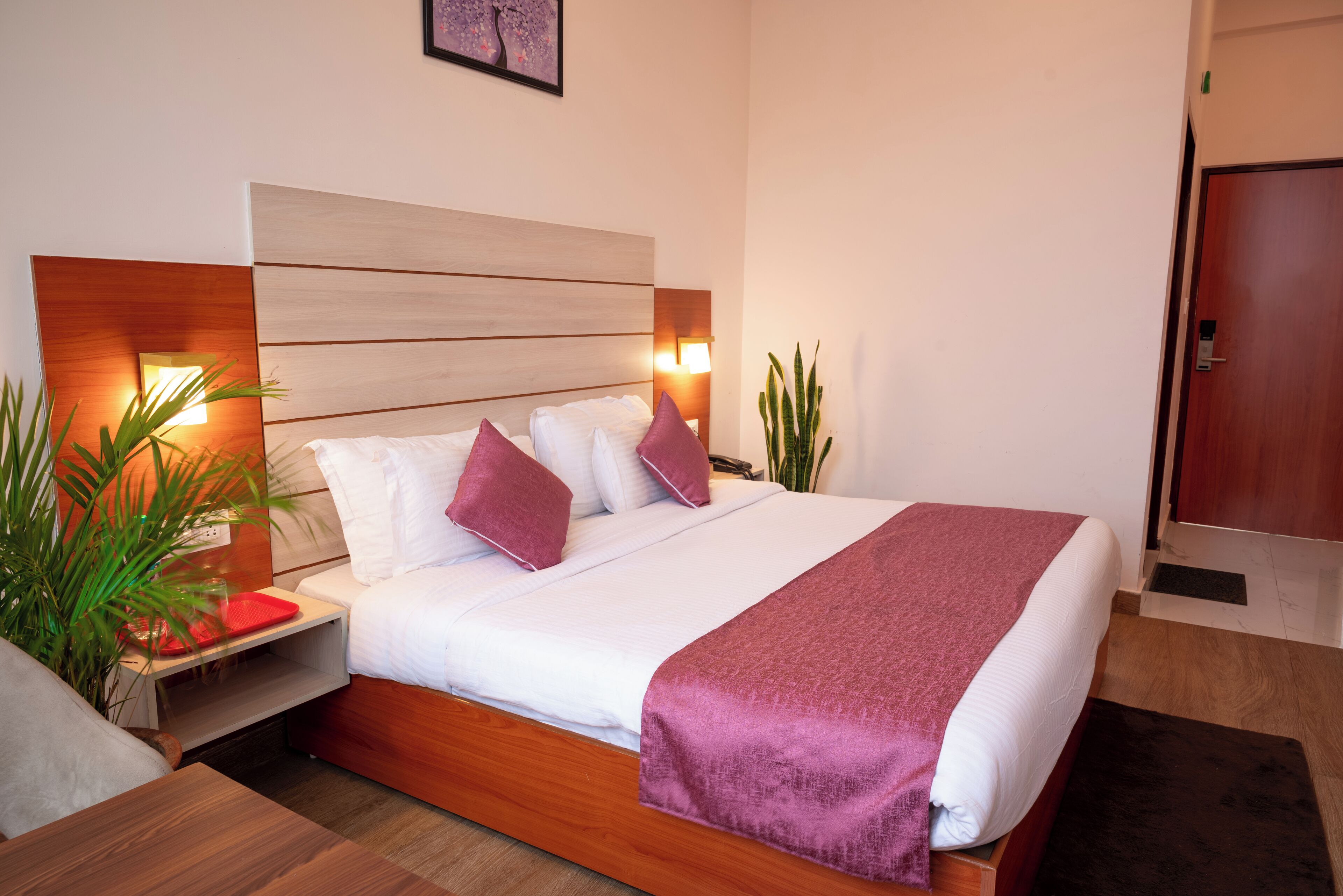Una habitación de hotel moderna con tablero de cabecera de madera, una cama grande con ropa de cama blanca y morada, una mesita de noche con bandeja roja y una mesita con lámpara.