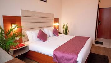 Una habitación de hotel moderna con tablero de cabecera de madera, una cama grande con ropa de cama blanca y morada, una mesita de noche con bandeja roja y una mesita con lámpara.