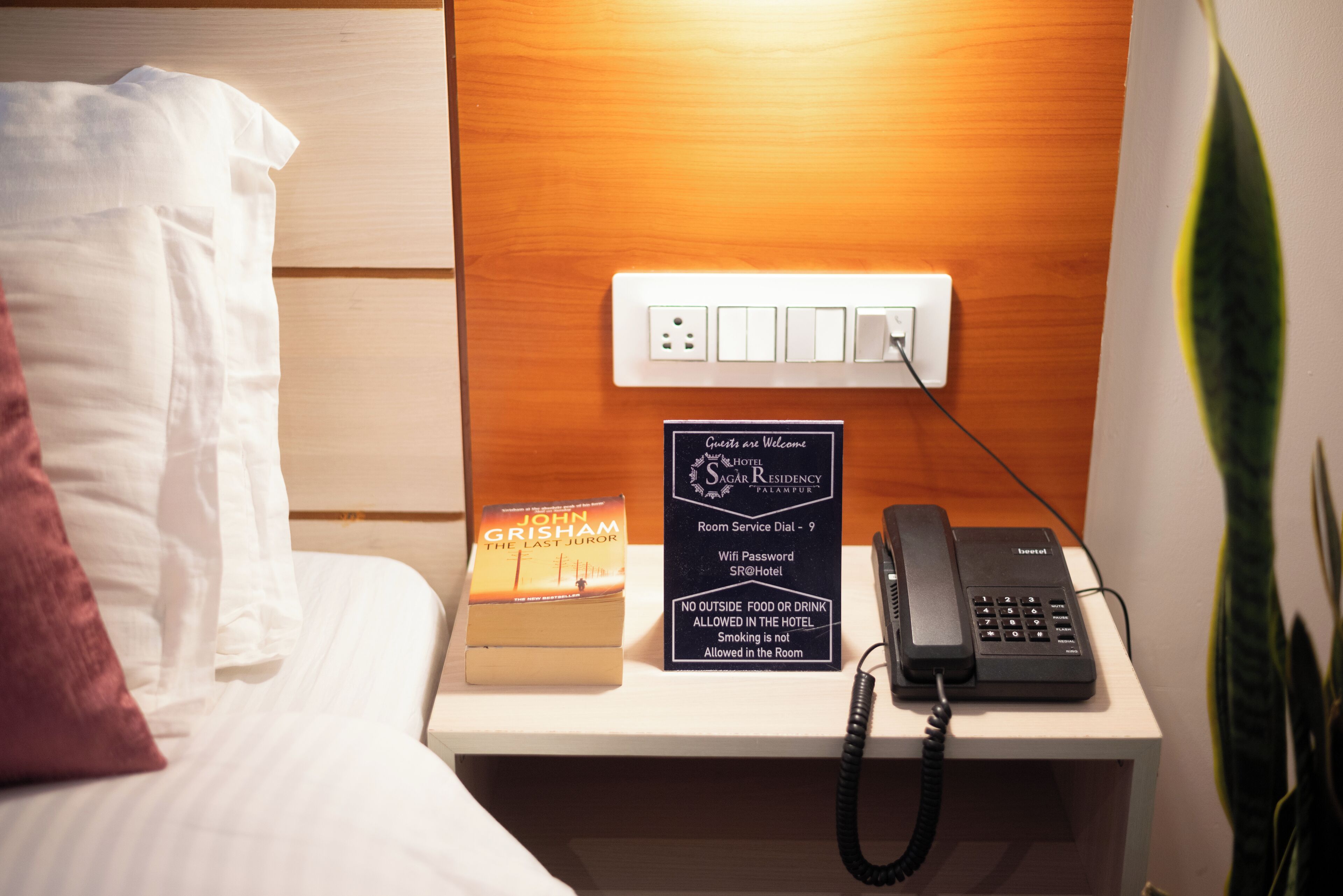 Habitación de hotel con una mesita de noche que incluye un teléfono, un cartel con información sobre servicio a la habitación y WiFi, y un libro titulado 