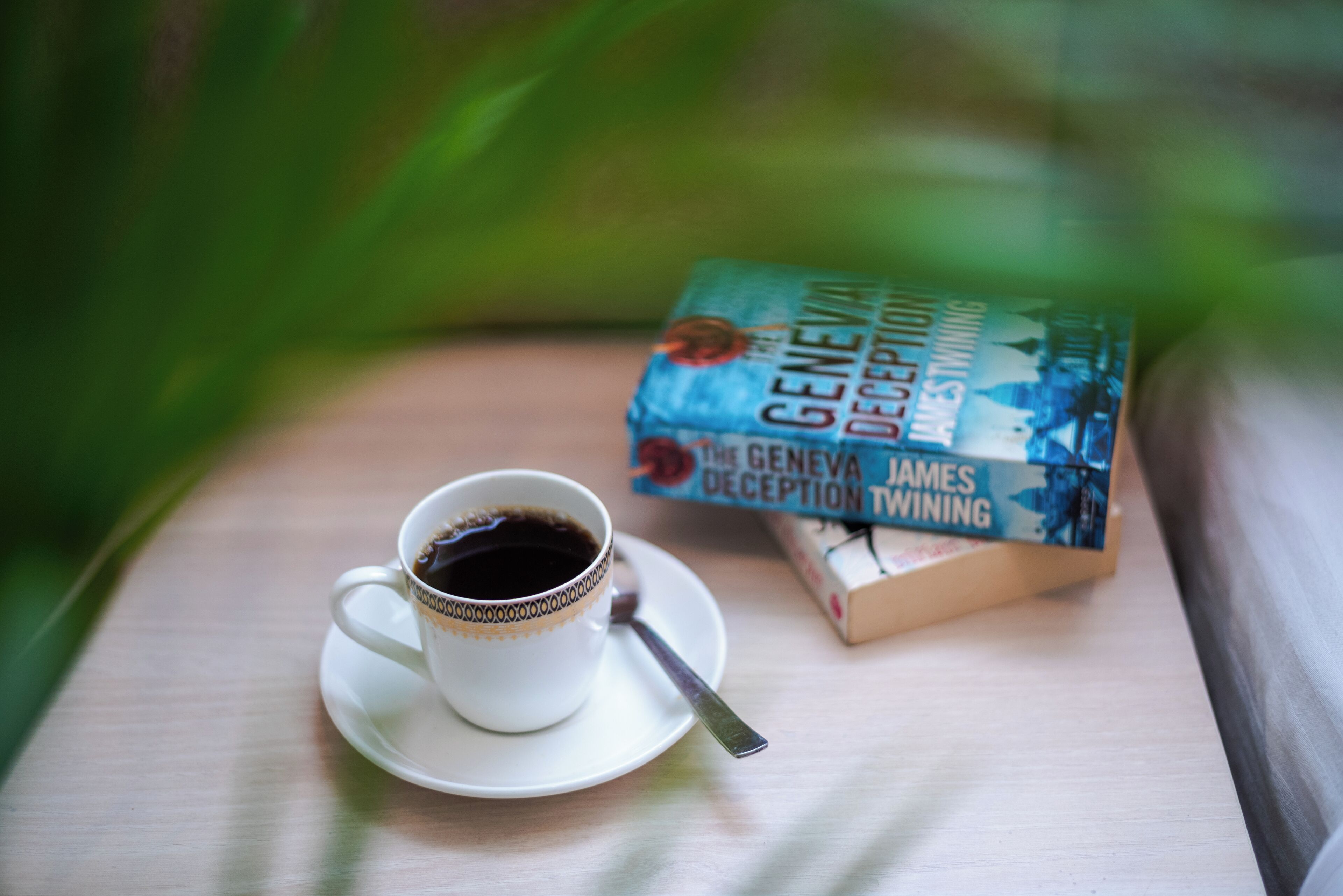A cup of coffee on a saucer with a spoon, next to a book titled 'The Geneva Deception' by James T. Winning.