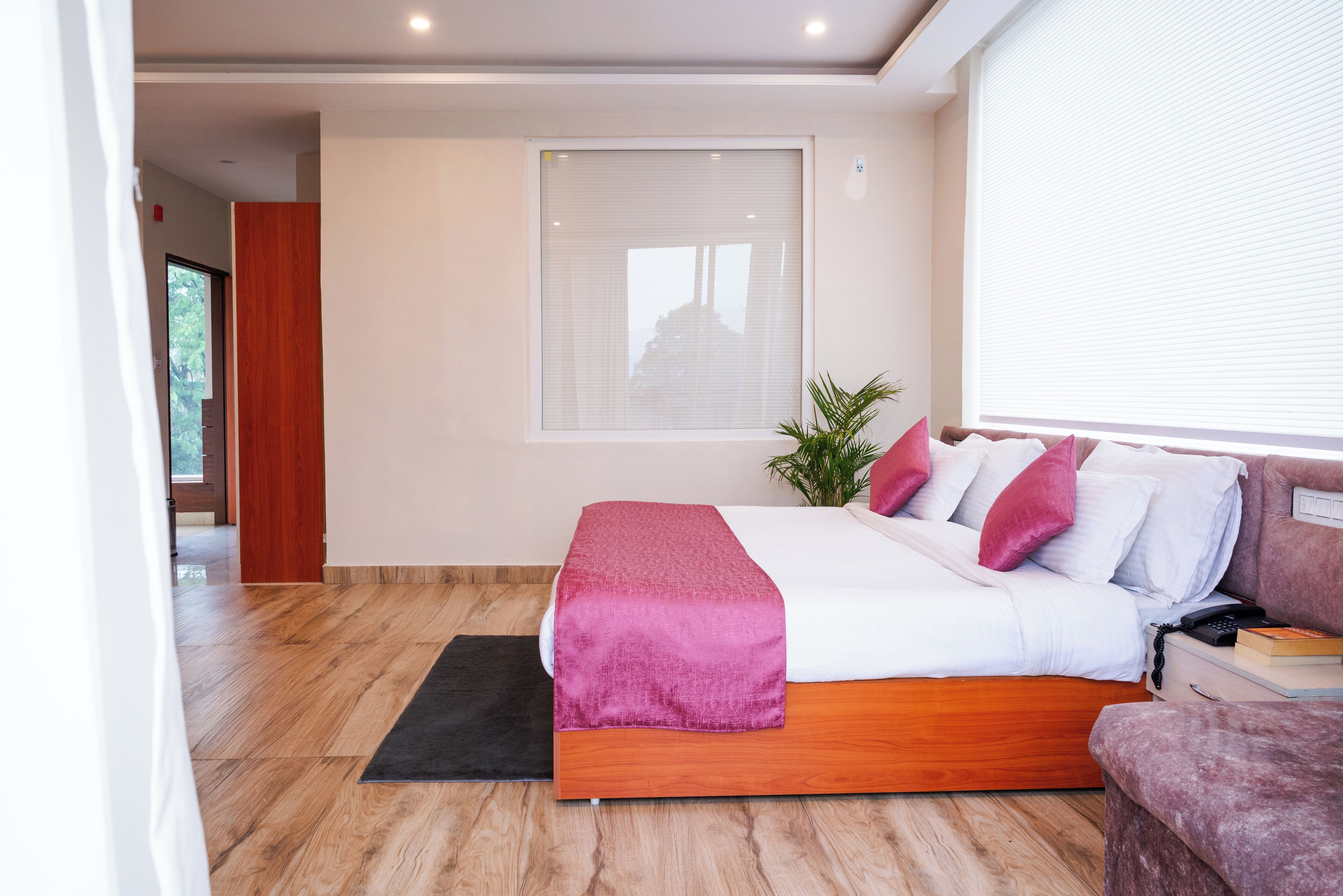 Une chambre moderne avec un plancher en bois, un grand lit recouvert d’une couverture rose, du linge de lit blanc et un mur d’accent rouge.