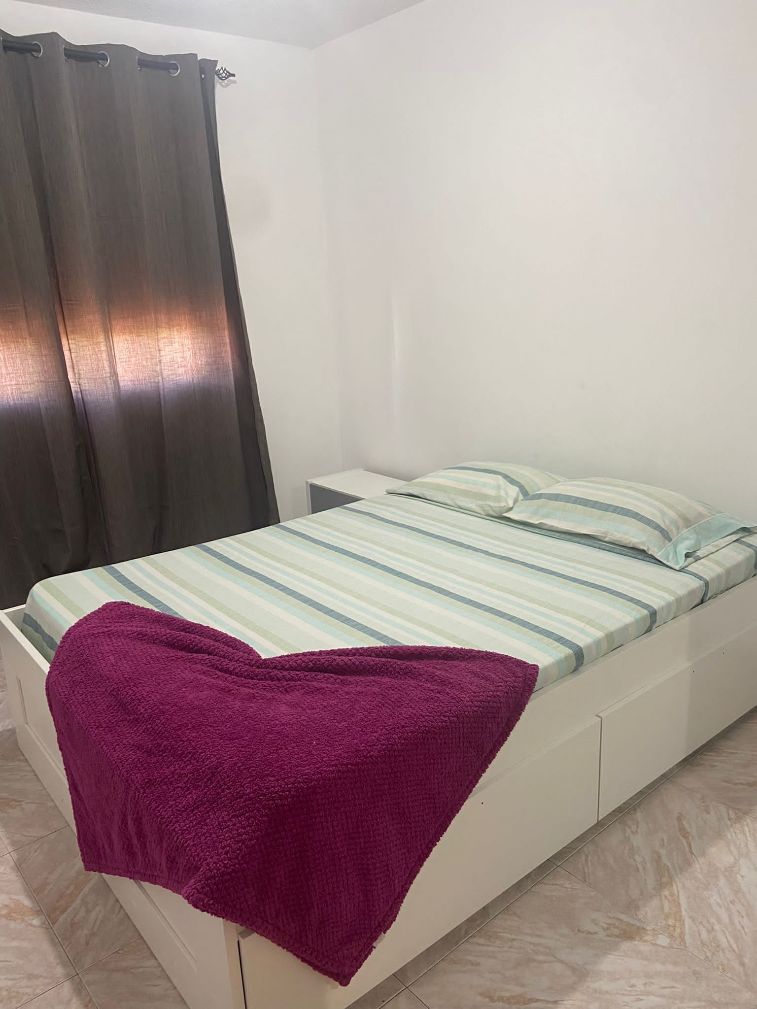 A single-bed room with a striped bedspread, a purple throw blanket, and a curtain.