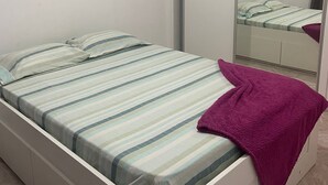 Een enkel bed met een gestreept beddekleed en een paarse deken, een grote spiegel en een witte kledingkast.