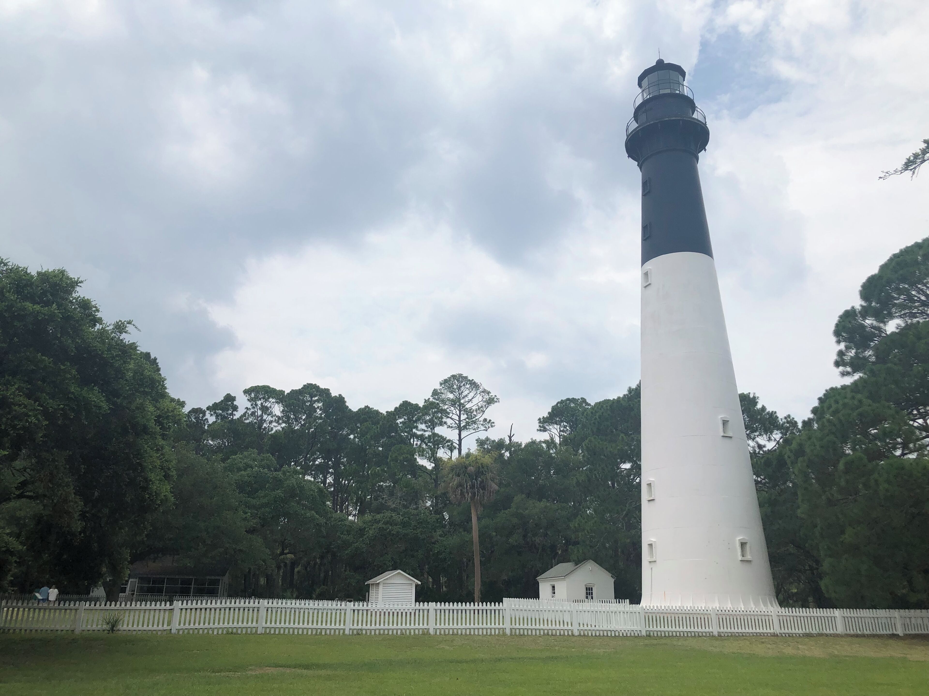 Un grand phare blanc, surmonté d’une partie noire, entouré d’une clôture blanche en bois et d’arbres.