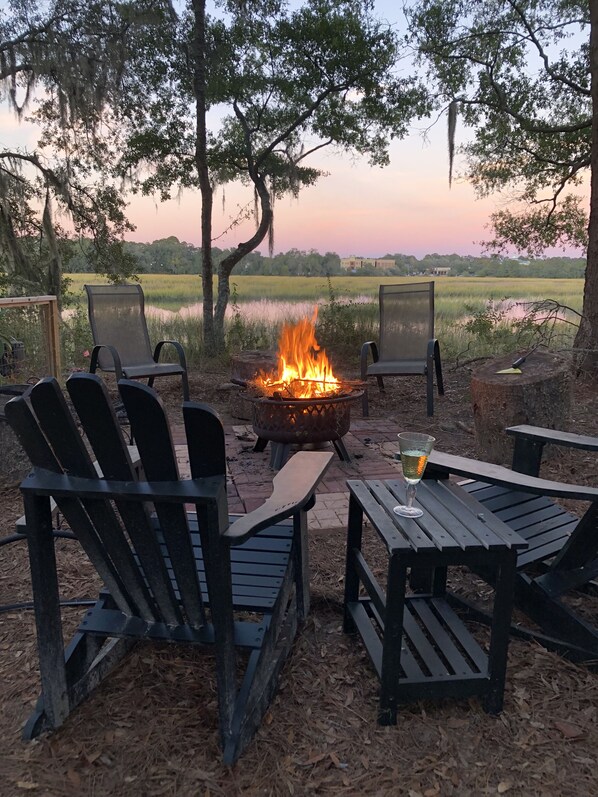 An outdoor seating area with chairs and tables, a fire pit, and a view of a field and building in the background.