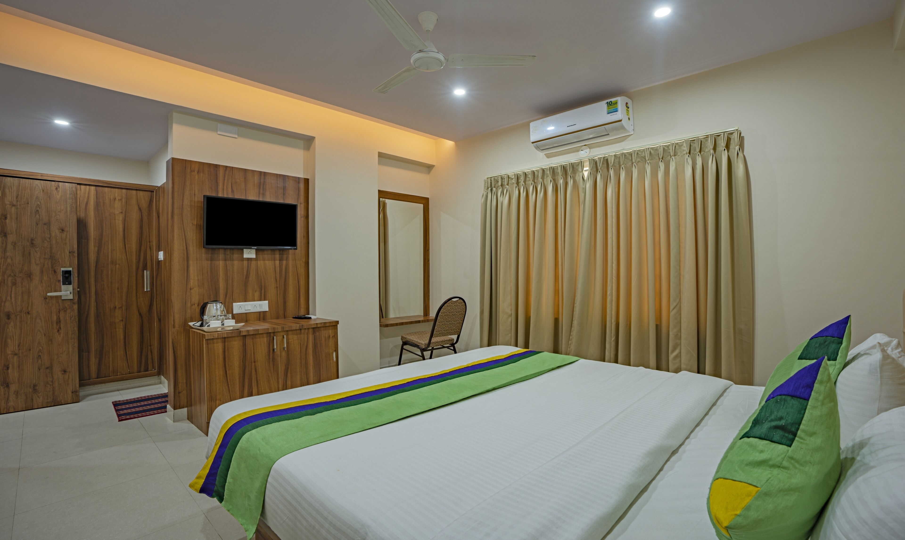 A hotel room with a bed, a desk with a television, an air conditioning unit, a chair, and curtains.