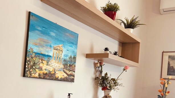 Ein Empfangsschalter mit einem Gemälde eines klassizistischen Gebäudes, Holzregale mit Pflanzen, einer Vase mit Blumen und einem Handdesinfektionsmittel.