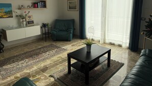 Ein Wohnzimmer mit einer Couch, einem türkisfarbenen Sessel, einem Couchtisch, einem Bücherregal und einem großen Fenster mit durchsichtigen Vorhängen.