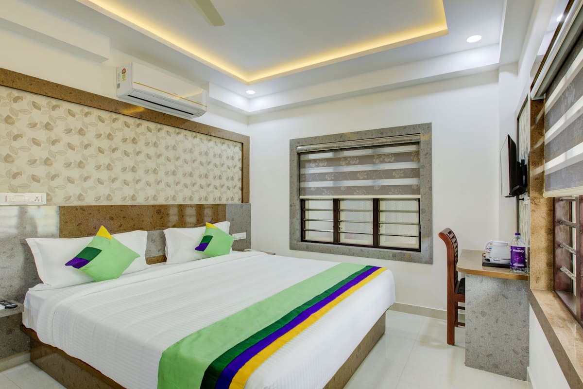 Quarto de hotel com cama, janela com persianas, ar condicionado, televisão e uma pequena mesa com uma garrafa.