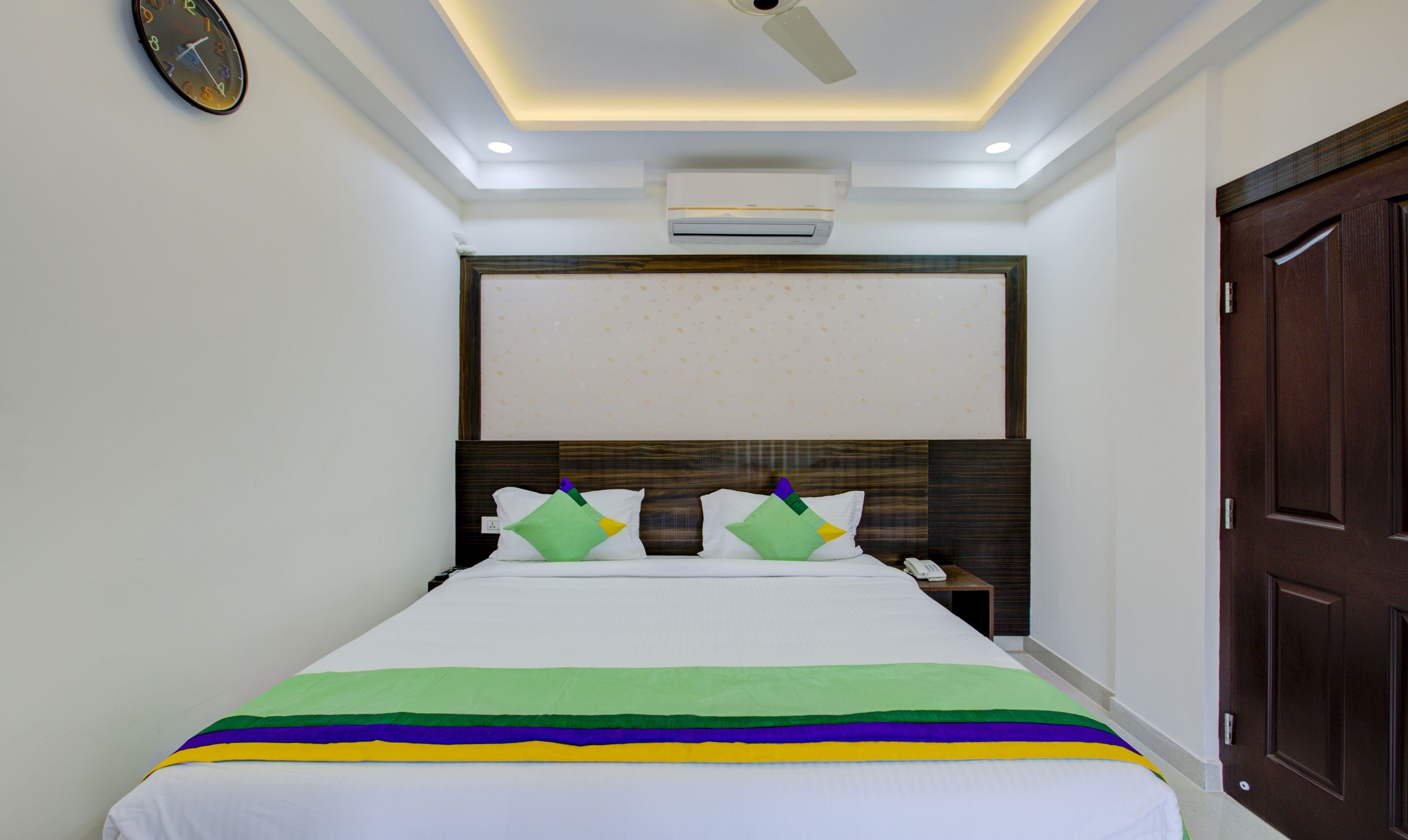 A hotel room with a large bed, a wooden headboard, a wall clock, an air conditioning unit, and a dark wooden door.