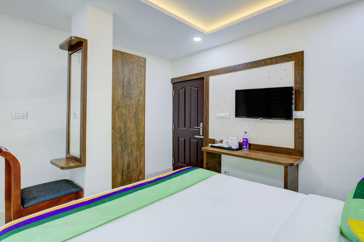 Quarto de hotel com cama, um banco de madeira, espelho, porta, TV e uma pequena mesa com uma garrafa.