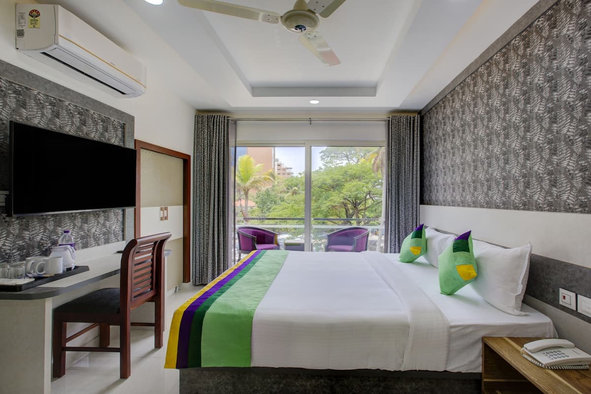 Quarto de hotel com cama, escrivaninha, cadeira, televisão e varanda com vista para árvores e edifícios.
