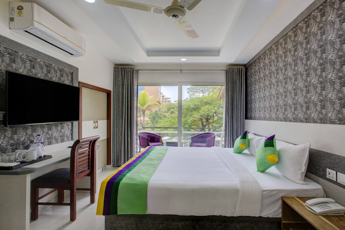 Quarto de hotel com cama, escrivaninha, cadeira, televisão, telefone e varanda com vista para árvores e edifícios.