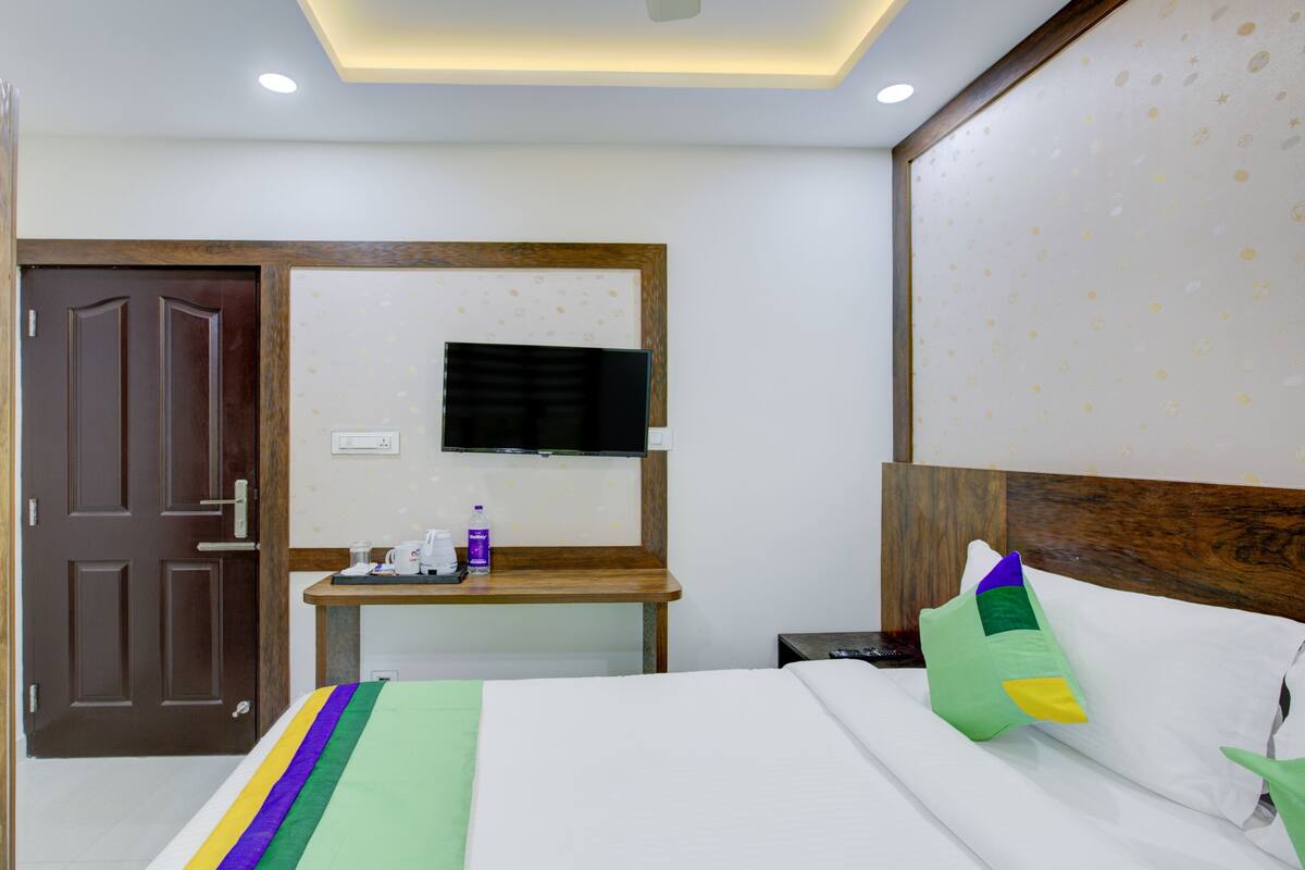 Quarto de hotel com uma cama, cabeceira de madeira, televisão fixada na parede, uma mesinha com uma garrafa e uma chávena, e uma porta com moldura de madeira.