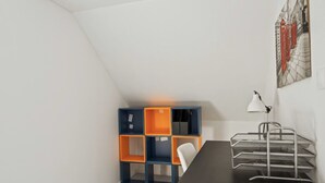 Une petite pièce bien éclairée, avec un plafond en pente, un bureau noir avec une lampe et une étagère colorée adossée au mur.