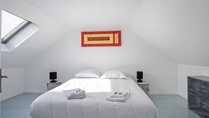 Une chambre moderne avec une lucarne, un grand lit recouvert de linge de maison blanc, deux tables de chevet équipées de haut-parleurs et une œuvre d’art encadrée en rouge accrochée au mur.