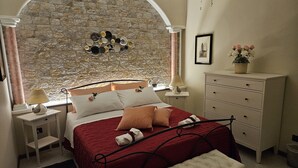 A bedroom with a stone wall, a bed with a red bedspread, a bench, a dresser, and a nightstand with a lamp.