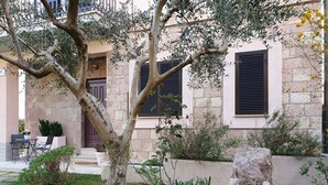 A stone building with two windows and a door, surrounded by greenery and a small outdoor seating area.