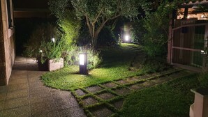 A nighttime garden with a paved pathway, illuminated by several lights, surrounded by greenery and a building in the background.