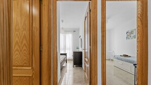 A hallway with wooden doors leading to a bedroom with a bed, bedside table, and a small desk with a chair. - Homely Lanzarote (Arrecife)