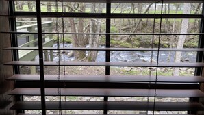 Ein Fenster mit horizontalen Jalousien, das Blick auf eine bewaldete Landschaft mit einem Bach und einer Holzterrasse bietet.