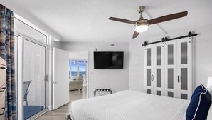 Un dormitorio con una cama, ventilador de techo, televisor y puerta corrediza que da a un balcón.