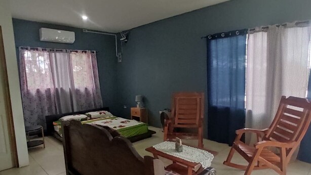 Una habitación con una cama, dos mecedoras, una mesita con una lámpara y una ventana con cortinas.
