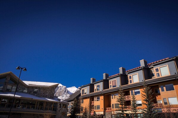 A multi-story building with a stone facade and a balcony, set against a backdrop of snow-capped mountains under a clear blue sky.