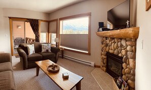 A cozy living room with a stone fireplace, a large window with blinds, a sofa with patterned cushions, and a television mounted above the fireplace.