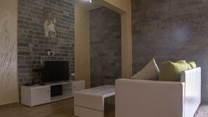 Una sala de estar con pared de ladrillo, un televisor de pantalla plana sobre un mueble blanco, un sofá blanco con un cojín floral y suelo de madera.