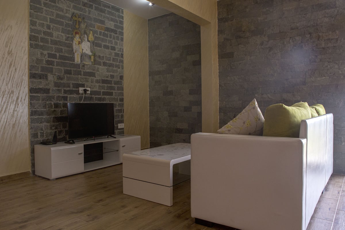 A living room with a brick wall, a flat-screen TV on a white cabinet, a white sofa with a floral cushion, and a wooden floor.