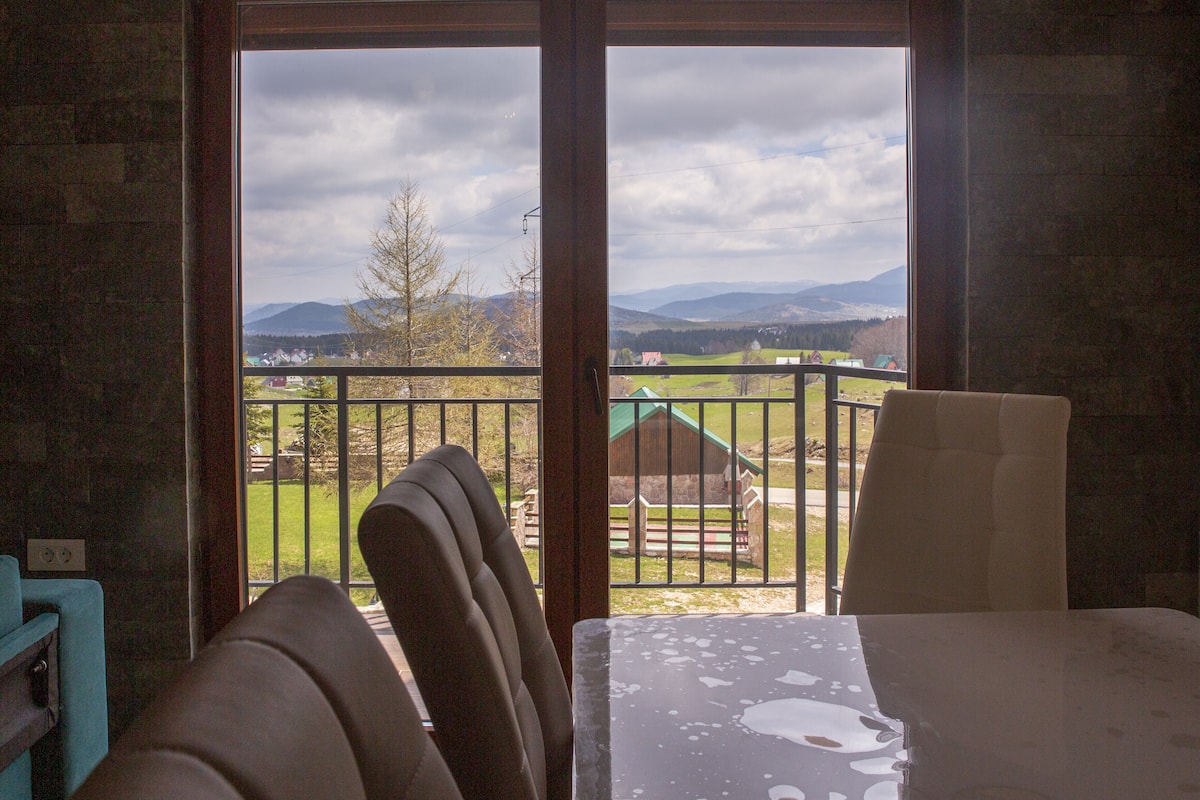 A room with a view of a green landscape, a balcony with a railing, and a table with a chair.